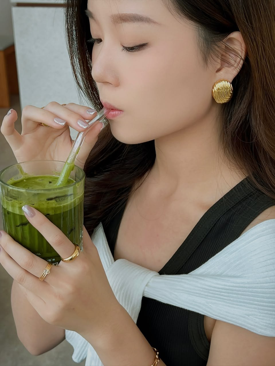 A close-up of Huan Min enjoying a matcha drink, with gold earrings and a white sweater.